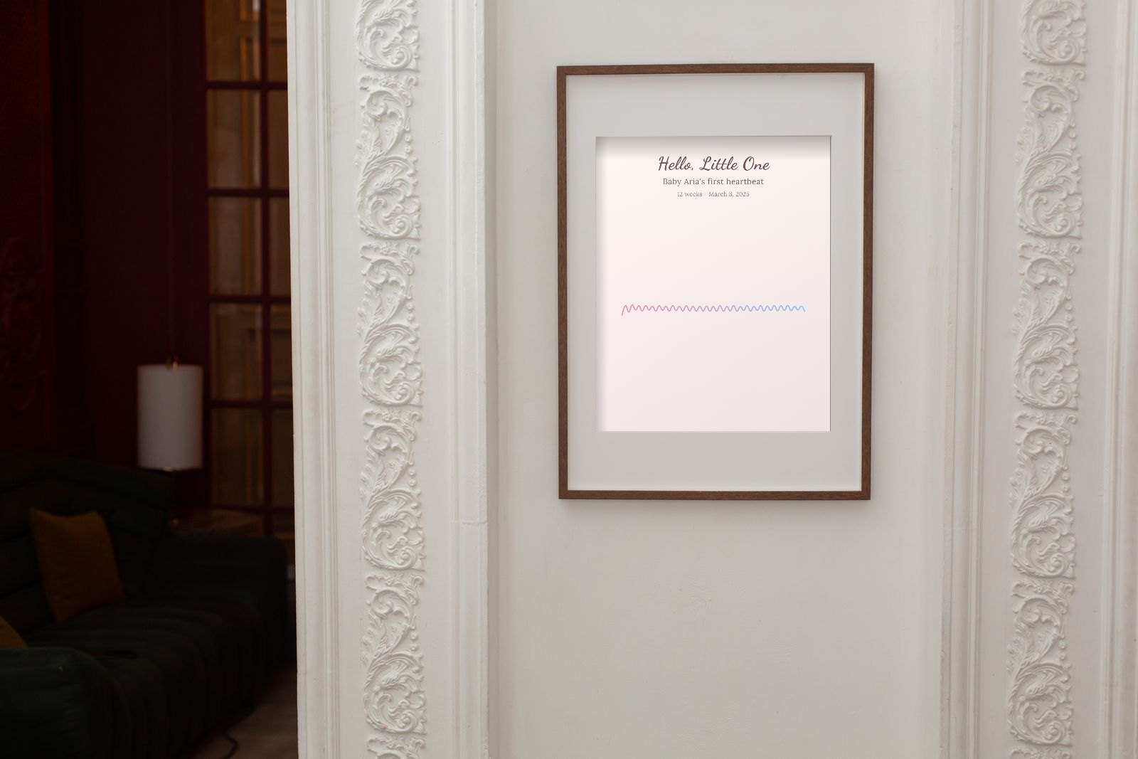 A wooden frame on a white panelled wall holding a pale pink print titled Hello, Little One with a thin pastel heartbeat line
