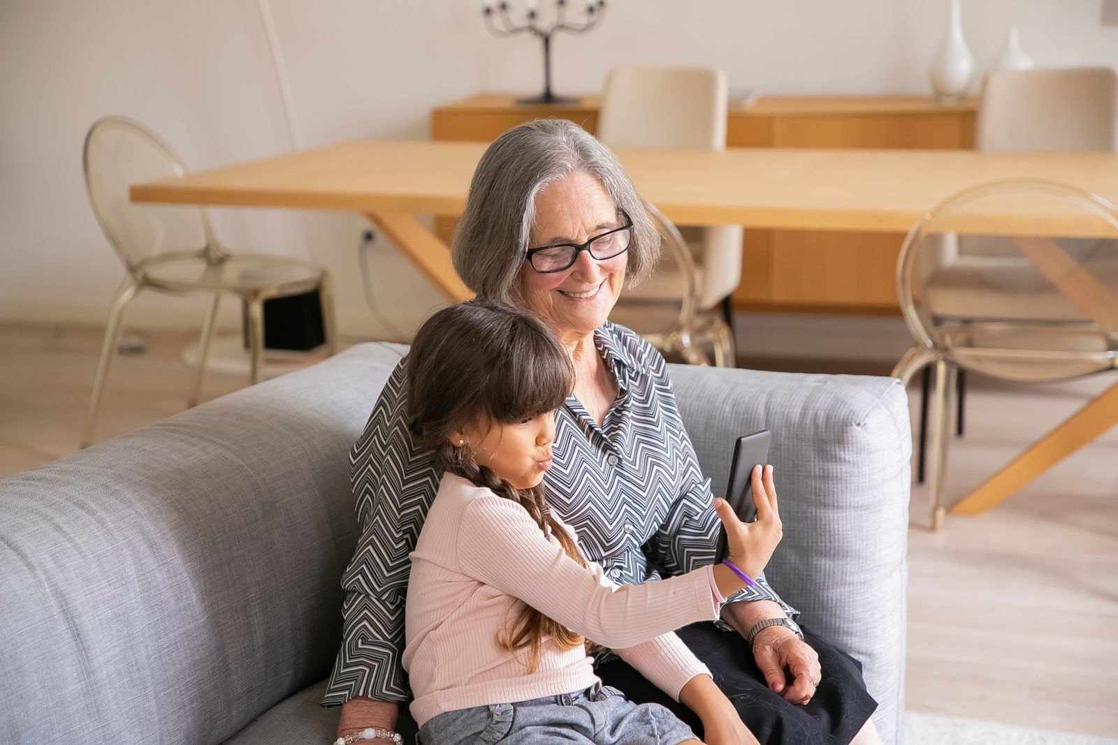 A grandmother and granddaughter smiling at a smartphone together on a sofa